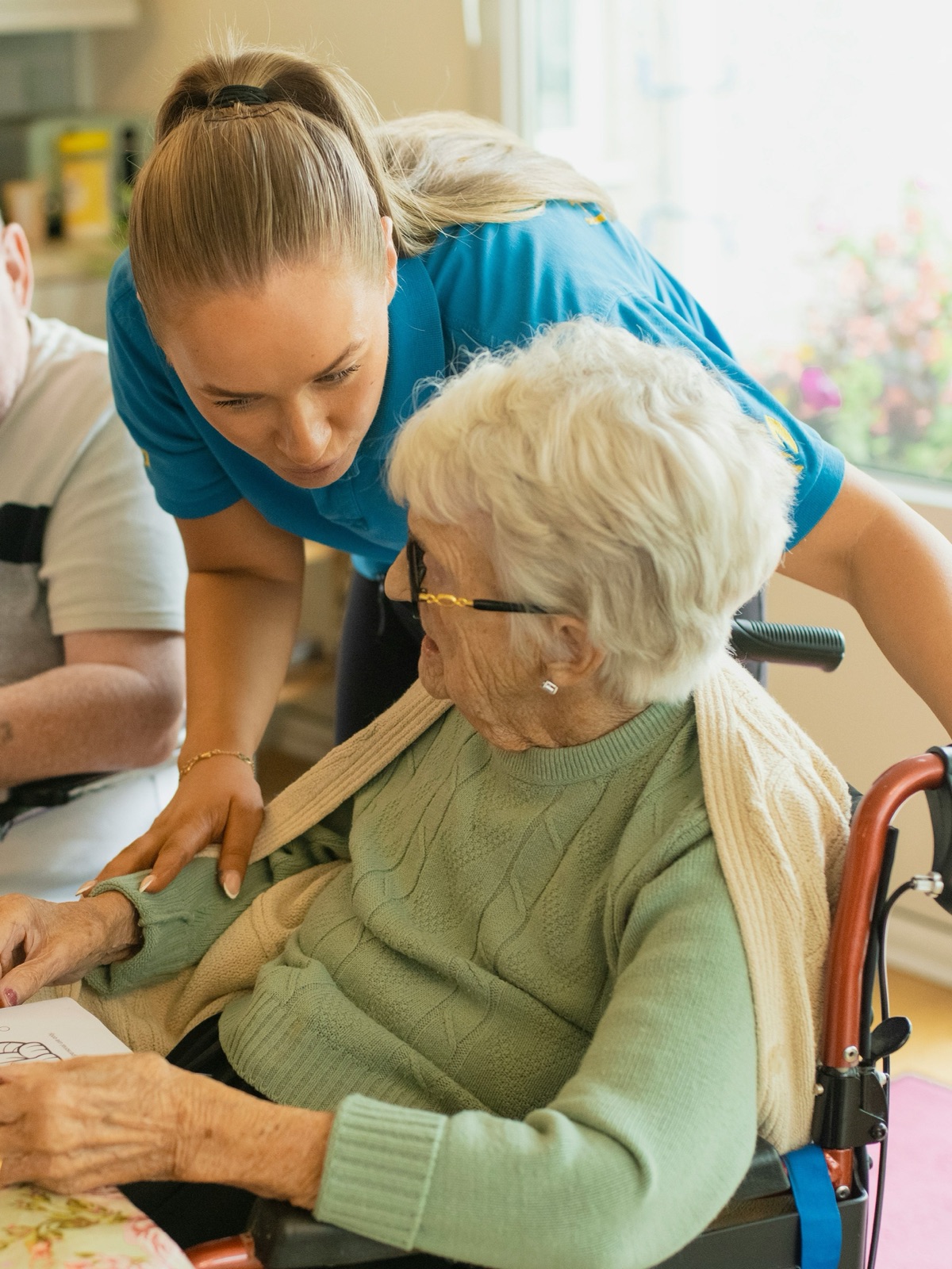 A support worker leaning in to help two residents at a group-home kitchen table