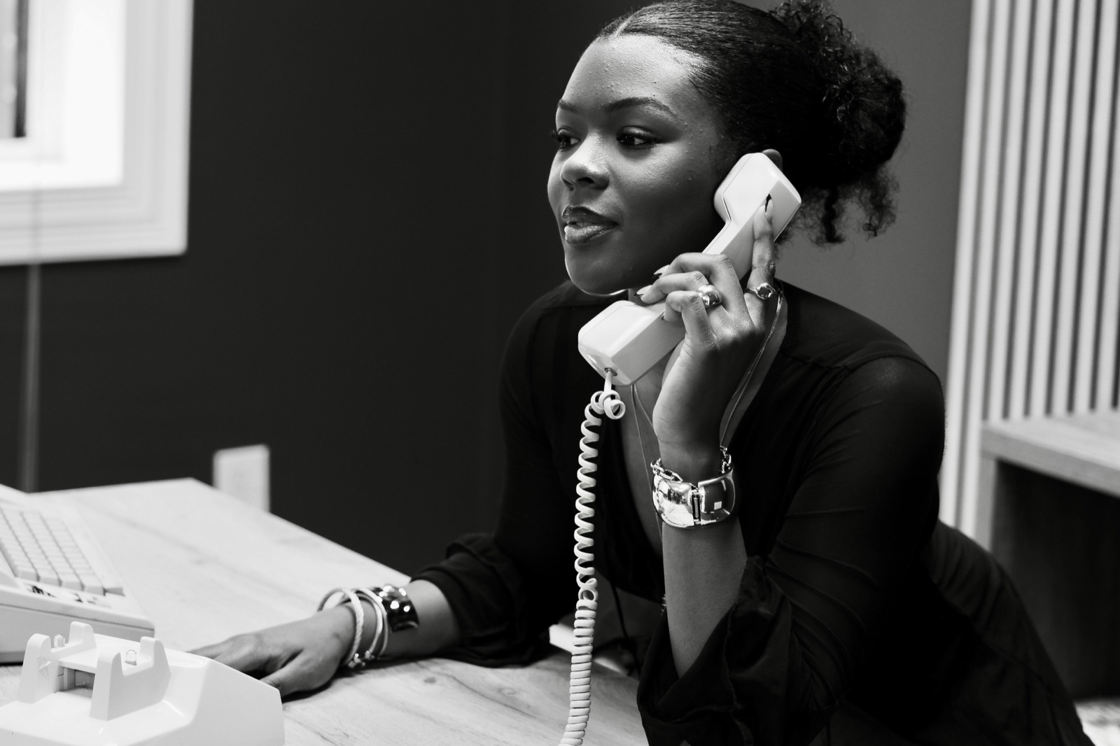 A woman at a desk holding a corded landline handset to her ear, in black and white.